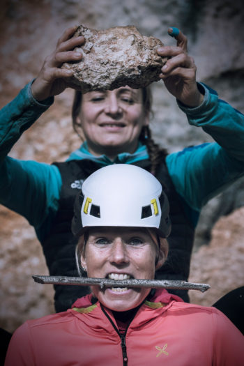 Eine Frau mit Kletterhelm über dem eine andere Frau bedrohlich einen Felsbrocken hält.