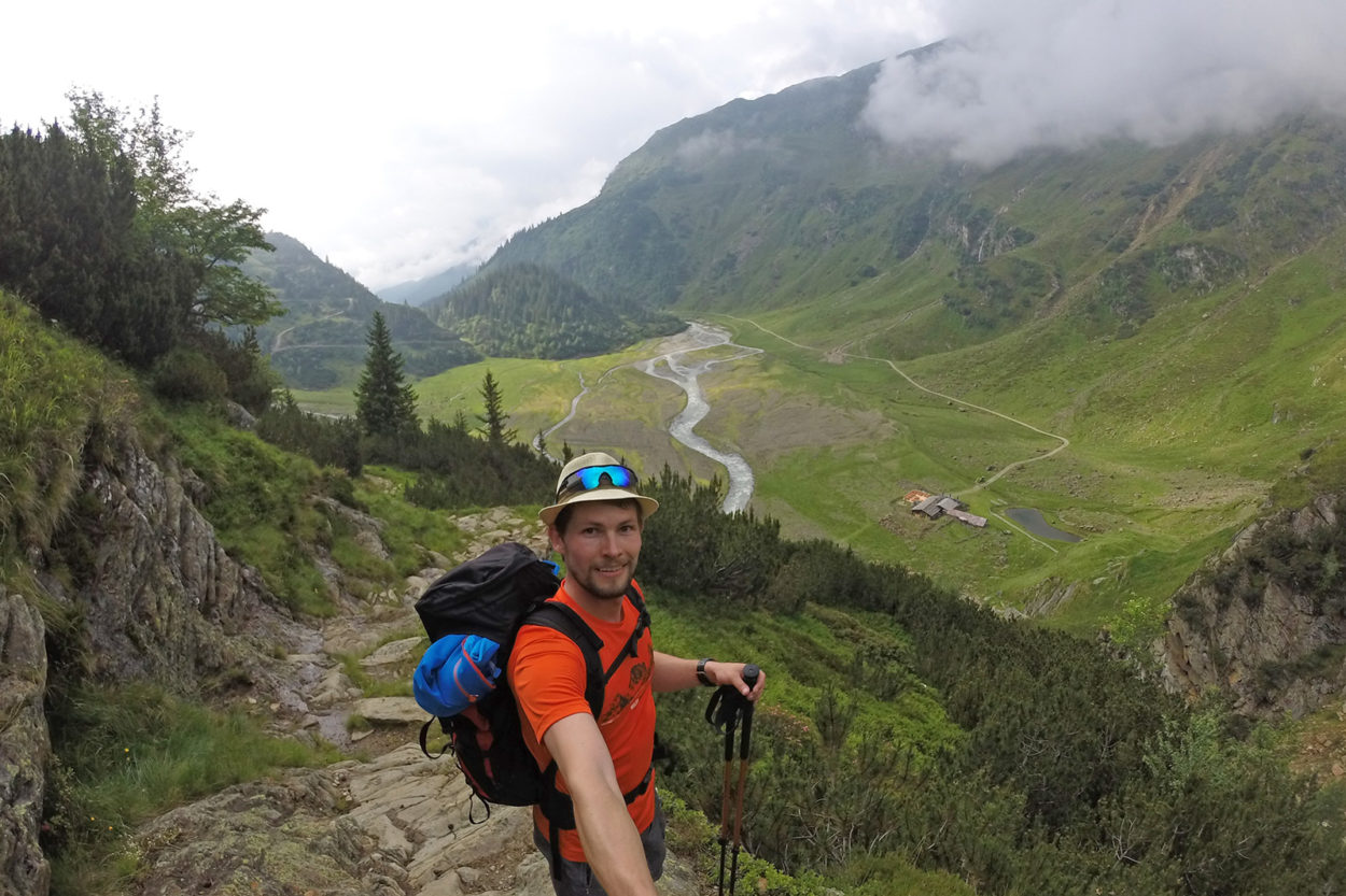 Ein Bergsteiger macht ein Selfie mit Blick auf eine gr&uuml;ne Hochalm, durch die sich ein Weg zieht.
