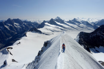 Zwei Bergsteiger laufen über einen schneebedeckten schmalen Grat, auf dem es zu beiden Seiten steil runtergeht. Im Hintergrund schneebedeckte Bergketten.